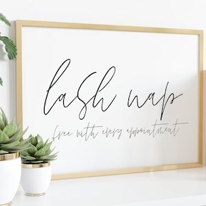 Puede incluir: Una impresión enmarcada con borde dorado muestra las palabras "lash nap" en escritura negra, con el texto "free with every appointment" debajo. La impresión está sobre una superficie blanca, con suculentas en macetas y vegetación en el fondo.