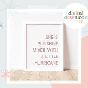 Puede incluir: Una impresión enmarcada en blanco con el texto "She is sunshine mixed with a little hurricane" en rosa, gris y turquesa. La impresión está sobre un suelo de madera clara.