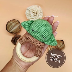 Könnte beinhalten: Ein grünes Häkelspielzeug Baby Yoda mit einem braunen Häkelschal, der an einem Holz-Beißring befestigt ist. Der Ring trägt die Gravur "Crocheteandotienda". Der Text "ENGLISH ESPAÑOL" ist in der unteren rechten Ecke des Bildes zu sehen.