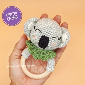 Puede incluir: Un sonajero de koala tejido a crochet con un collar tejido a crochet verde y un anillo de madera. El koala tiene una nariz negra y ojos bordados.