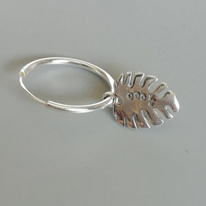 May include: Silver hoop earring with a silver leaf charm. The leaf charm has a textured surface and a small stamp on the back.