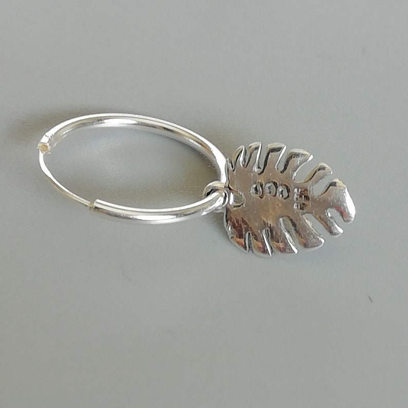 May include: Silver hoop earring with a silver leaf charm. The leaf charm has a textured surface and a small stamp on the back.
