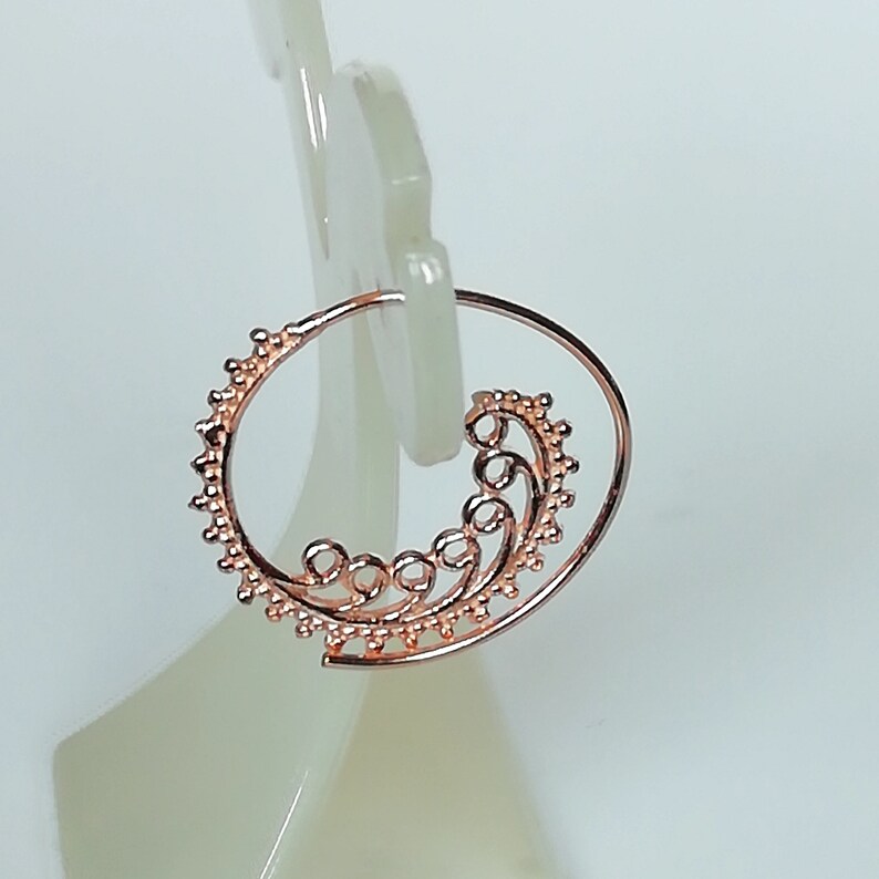 May include: Rose gold hoop earrings with a delicate, swirling design. The earrings are made of metal and have a circular shape.