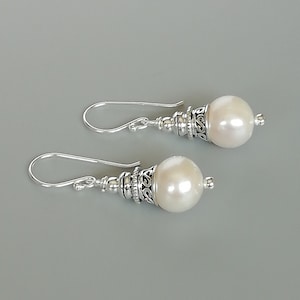 Puede incluir: Un par de pendientes elegantes con grandes perlas blancas cremosas. Las perlas están adornadas con intrincados detalles de metal plateado y cuelgan de sencillos ganchos curvos. Fondo gris suave.