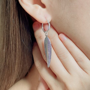 Puede incluir: Pendientes de aro de plata con un dije de pluma que cuelga de la parte inferior. El dije de pluma tiene un dise&ntilde;o texturizado.