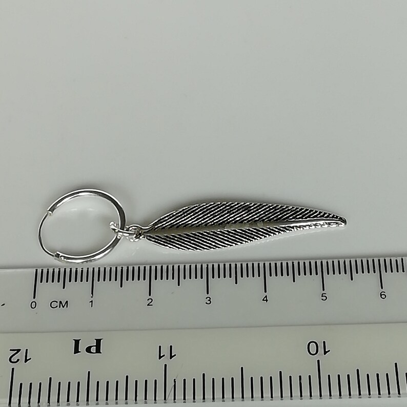 Puede incluir: Pendiente de aro de plata con un dije de pluma. El pendiente mide aproximadamente 5 cm de largo.