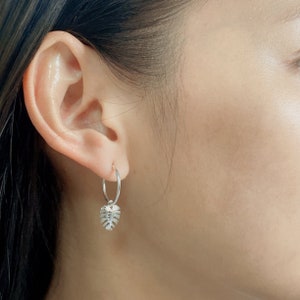 May include: Silver hoop earrings with a small, silver, leaf-shaped charm hanging from the bottom.