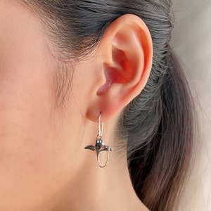 Puede incluir: Pendientes de plata con un pequeño encanto de raya manta detallada. Los pendientes son usados por una persona con cabello largo y oscuro.