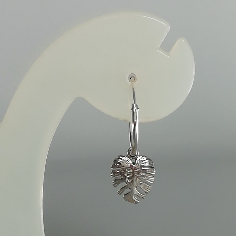 May include: Silver hoop earrings with a small, detailed leaf charm hanging from the bottom.