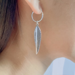 Puede incluir: Pendientes de aro de plata con un dije de pluma que cuelga de la parte inferior. El dije de pluma tiene un dise&ntilde;o detallado.
