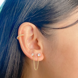 Peut inclure: Boucles d'oreilles en chaîne dorée avec de petites pierres claires. La chaîne relie deux clous d'oreille.