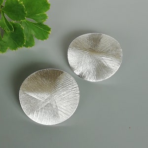 Könnte beinhalten: Ein Paar silberne Ohrringe mit einem runden, strukturierten Design. Die Ohrringe haben eine gebürstete Oberfläche und eine leicht erhöhte Mitte.