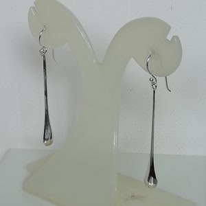 May include: A pair of silver teardrop earrings hanging from a display stand. The earrings are long and slender, with a smooth, rounded teardrop shape at the bottom.