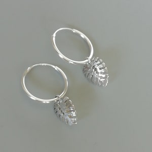 May include: A pair of silver hoop earrings with a dangling leaf charm. The leaves are detailed and have a cutout design.