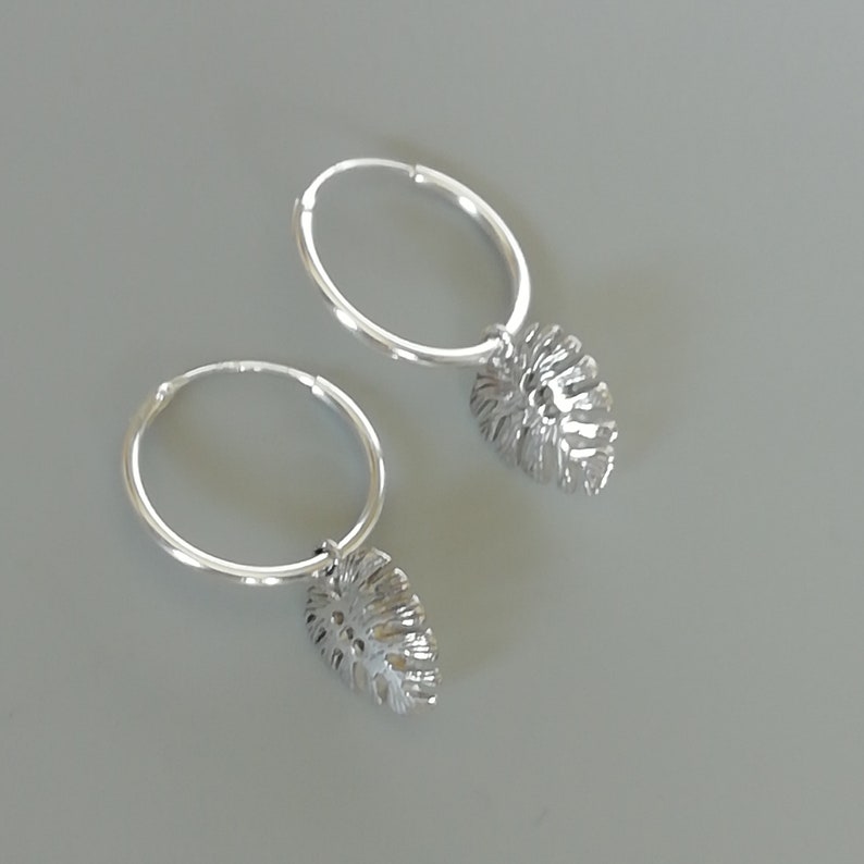 May include: A pair of silver hoop earrings with a dangling leaf charm. The leaves are detailed and have a cutout design.