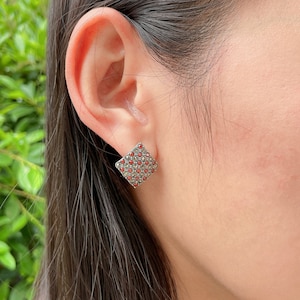May include: Square stud earrings with a silver-tone metal setting. The earrings are covered in small, round, red and silver-colored stones. The earrings are shown on an ear, with the person's hair and skin visible.