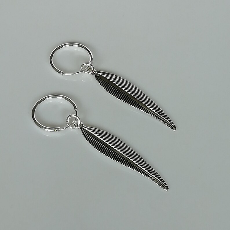 Puede incluir: Un par de pendientes de aro de plata con colgantes de plumas. Las plumas tienen un dise&ntilde;o texturizado.