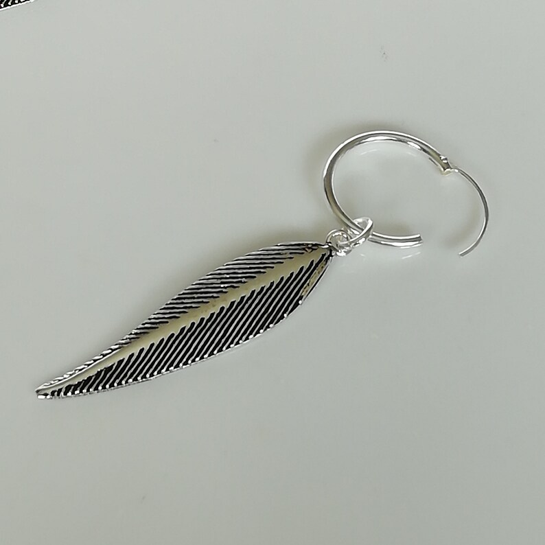 Puede incluir: Pendiente de aro de plata con un dije de pluma. La pluma est&aacute; detallada con l&iacute;neas y tiene un acabado plateado.