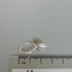 May include: Silver hoop earrings with a small, silver leaf charm hanging from the hoop. The earrings are shown next to a ruler, measuring approximately 2 cm in diameter.