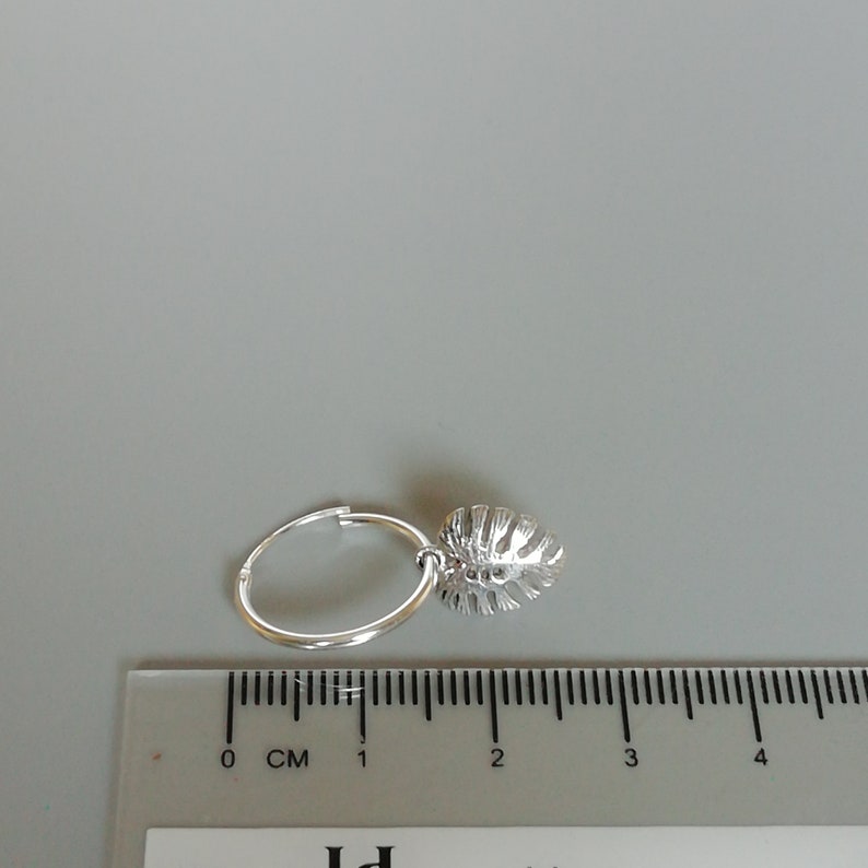 May include: Silver hoop earrings with a small, silver leaf charm hanging from the hoop. The earrings are shown next to a ruler, measuring approximately 2 cm in diameter.