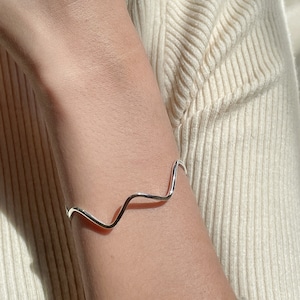 May include: A silver wavy bangle bracelet on a person's wrist.