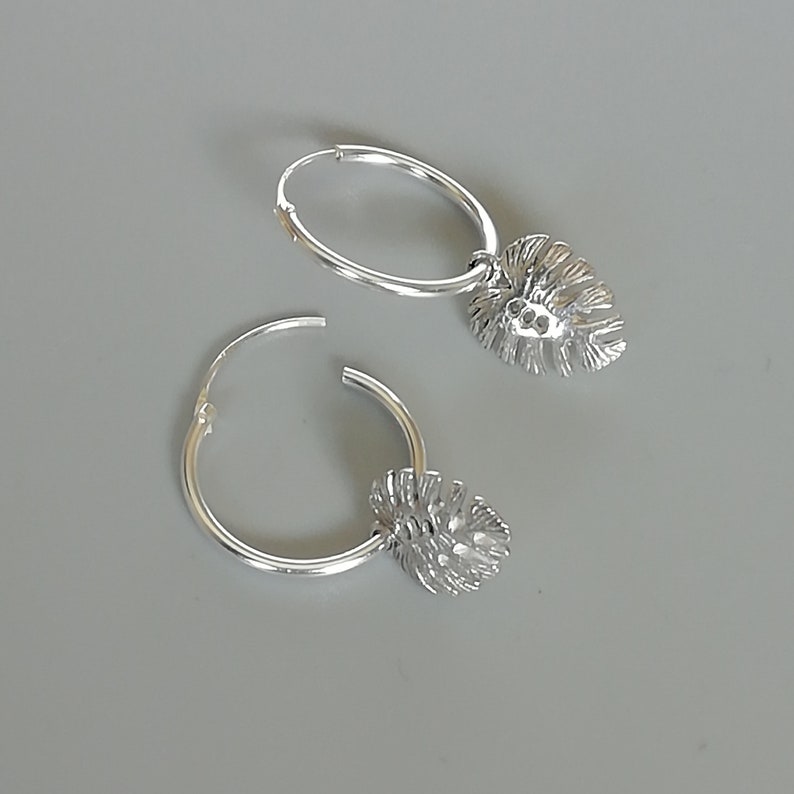 May include: A pair of silver hoop earrings with a leaf-shaped charm hanging from each hoop.
