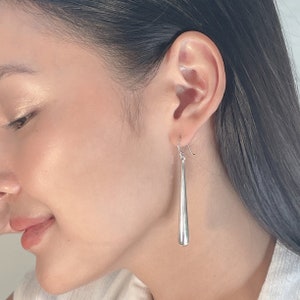 Puede incluir: Pendientes plateados con forma de lágrima. Estos pendientes colgantes tienen un diseño elegante y alargado en forma de lágrima. Los pendientes están hechos de un metal brillante y reflectante, y los ganchos son visibles. Miden aproximadamente 7.6 cm de largo.