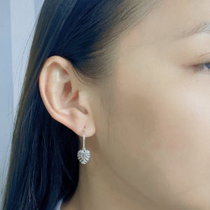 May include: Silver hoop earrings with a small, detailed leaf charm hanging from the bottom.