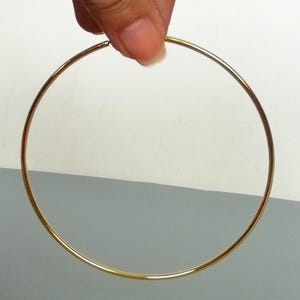Könnte beinhalten: Ein goldfarbener Creolen-Ohrring. Der Ohrring hat ein einfaches, kreisförmiges Design mit glatter, polierter Oberfläche. Der Ohrring wird von den Fingern einer Person gehalten.