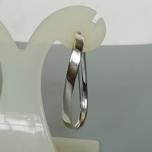 May include: A single silver-colored hoop earring with a polished finish. The earring has a teardrop shape and is displayed on a white earring stand against a gray background. The earring is a simple, elegant design.