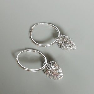 May include: A pair of silver hoop earrings with a leaf charm hanging from each hoop.