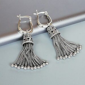 May include: A pair of silver earrings with a tassel design. The earrings are made of a silver chain with a small, round, silver bead at the end of each chain. The earrings are attached to a silver hoop.