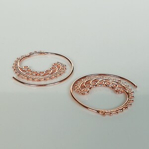 May include: A pair of rose gold spiral hoop earrings with a delicate, intricate design. The earrings are made of thin wire and have a circular shape.