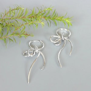May include: A pair of silver-toned bow earrings. Each earring features a bow design with looped details and long, curved streamers. The earrings are set against a gray background with a sprig of green foliage in the upper left corner.