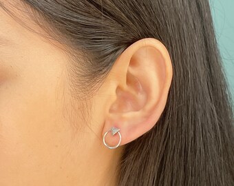 May include: A close-up of a silver earring on an ear. The earring features a small, open circle with a textured triangle design at the top. The earring is a stud style, and the metal appears to be polished.