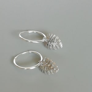 May include: A pair of silver hoop earrings with a dangling leaf charm. The leaf charm is detailed and has a textured surface.