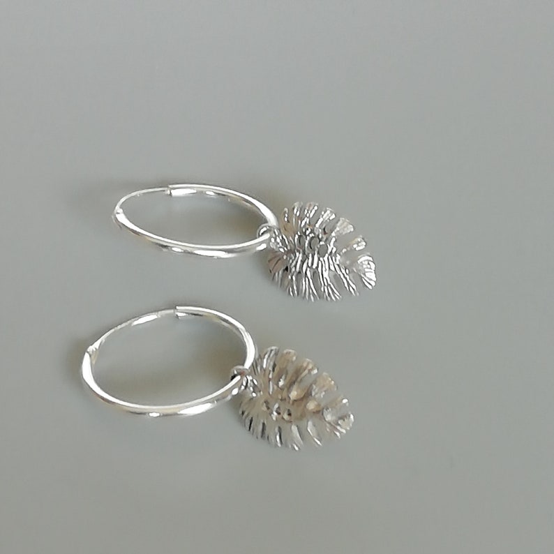 May include: A pair of silver hoop earrings with a dangling leaf charm. The leaf charm is detailed and has a textured surface.