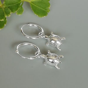 May include: A pair of silver hoop earrings with small turtle charms. The earrings are displayed on a gray surface with green foliage in the background. The turtle charms have detailed shell and flipper designs.