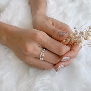 Może przedstawiać: Srebrny pierścionek o unikalnym, otwartym wzorze z prostokątnymi ogniwami. Pierścionek jest prezentowany na dłoni z brokatowym lakierem do paznokci, na miękkim, białym tle. Dłoń trzyma również mały bukiet suszonych kwiatów.