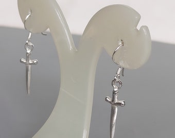 May include: A pair of silver earrings featuring a dagger design. The earrings are displayed on a white stand.
