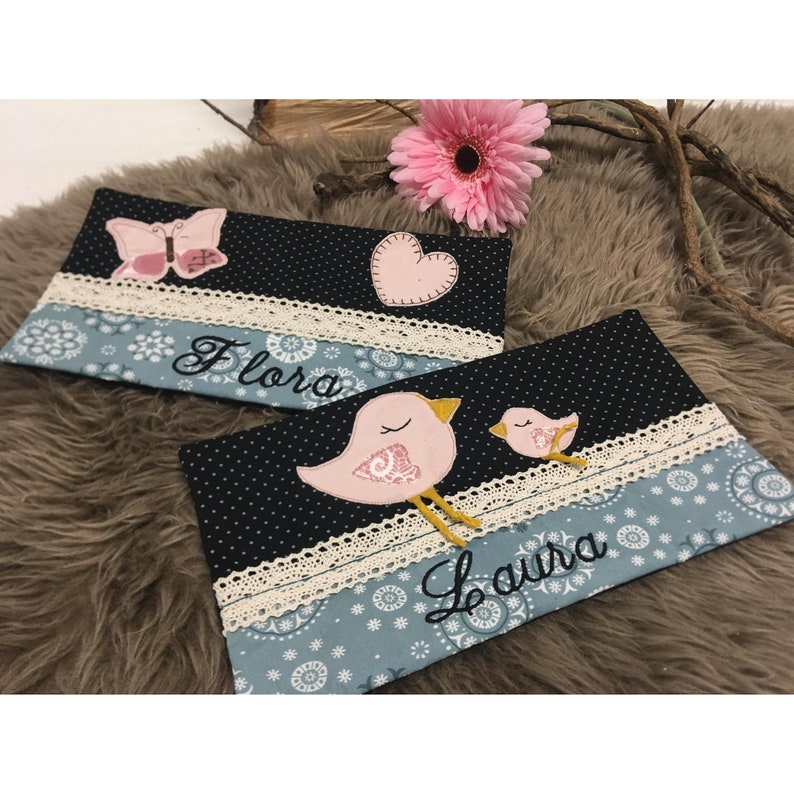 Op de afbeelding: Twee zwarte en blauwe stoffen zakjes met witte kantversiering. De zakjes zijn geborduurd met vogels, harten en vlinders. E&eacute;n zakje heeft de naam "Flora" geborduurd, en de andere heeft de naam "Laura" geborduurd.