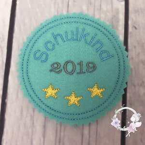 Pode incluir: Um c&iacute;rculo de feltro azul turquesa com o texto "Schulkind 2019" em bordado cinza. Tr&ecirc;s estrelas amarelas bordadas est&atilde;o na parte inferior do c&iacute;rculo.