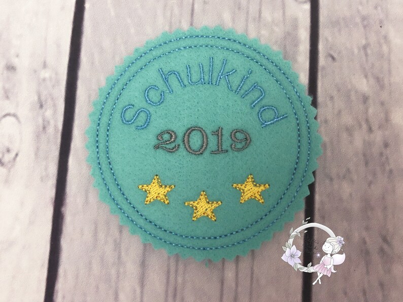 Pode incluir: Um c&iacute;rculo de feltro azul turquesa com o texto "Schulkind 2019" em bordado cinza. Tr&ecirc;s estrelas amarelas bordadas est&atilde;o na parte inferior do c&iacute;rculo.