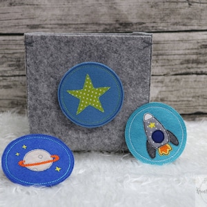 Könnte beinhalten: Drei Filzapplikationen mit Weltraumthema. Ein blauer Kreis mit einem gelben Stern, ein blauer Kreis mit einer Rakete und ein blauer Kreis mit einem Planeten.