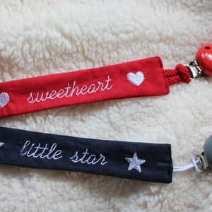 May include: Two fabric baby soothers with wooden beads. One is red with white hearts and the text "sweetheart" embroidered on it. The other is navy blue with white stars and the text "little star" embroidered on it.