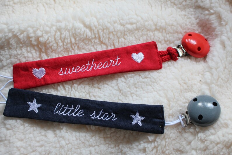 May include: Two fabric baby soothers with wooden beads. One is red with white hearts and the text "sweetheart" embroidered on it. The other is navy blue with white stars and the text "little star" embroidered on it.