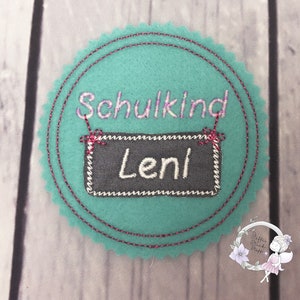 Pode incluir: Um c&iacute;rculo de feltro turquesa com uma borda costurada rosa. O c&iacute;rculo tem o texto "Schulkind" e um quadro-negro com o nome "Leni" escrito em branco.