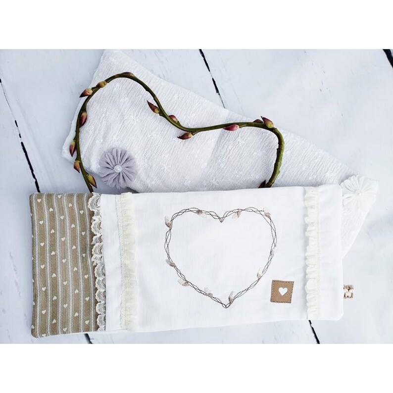 May include: A white fabric pouch with a heart-shaped embroidery design. The heart is outlined with a brown floral pattern. The pouch has a brown and white striped fabric border and a white lace trim.