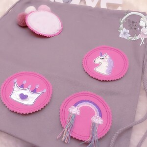 Puede incluir: Una bolsa de cordón gris con cuatro parches de fieltro rosa. Los parches representan un unicornio, una corona con un corazón, un arcoíris con nubes y un círculo en blanco. La bolsa tiene una etiqueta blanca con el texto "Stuff's Stick Stuff".