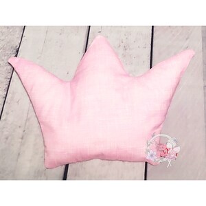 May include: A pink crown-shaped pillow made of soft fabric. The pillow is on a light wood surface.